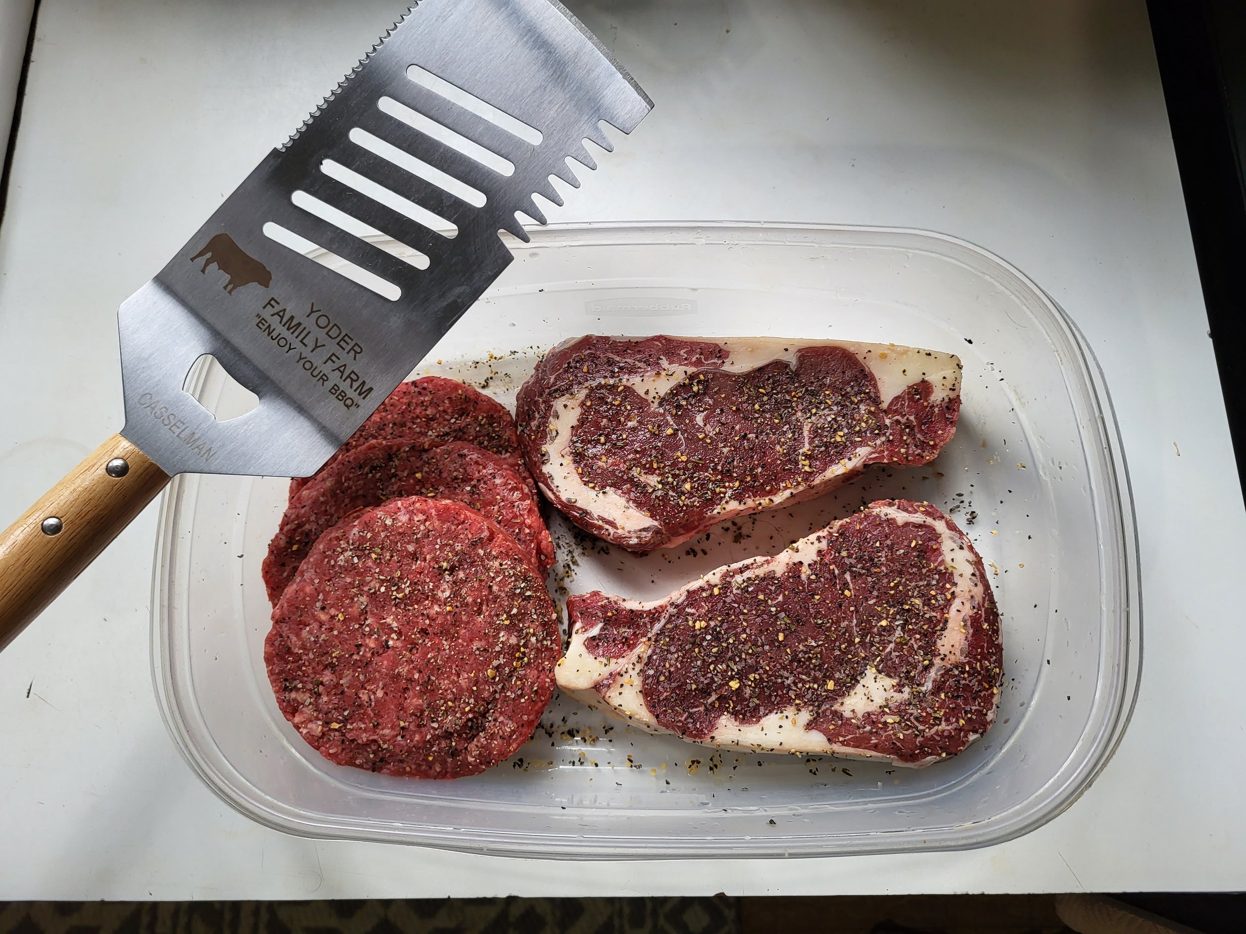 Raw beef patties and ribeye steaks seasoned with spices, placed in a clear container, accompanied by a stainless steel spatula with a wooden handle, ideal for grilling and barbecuing.