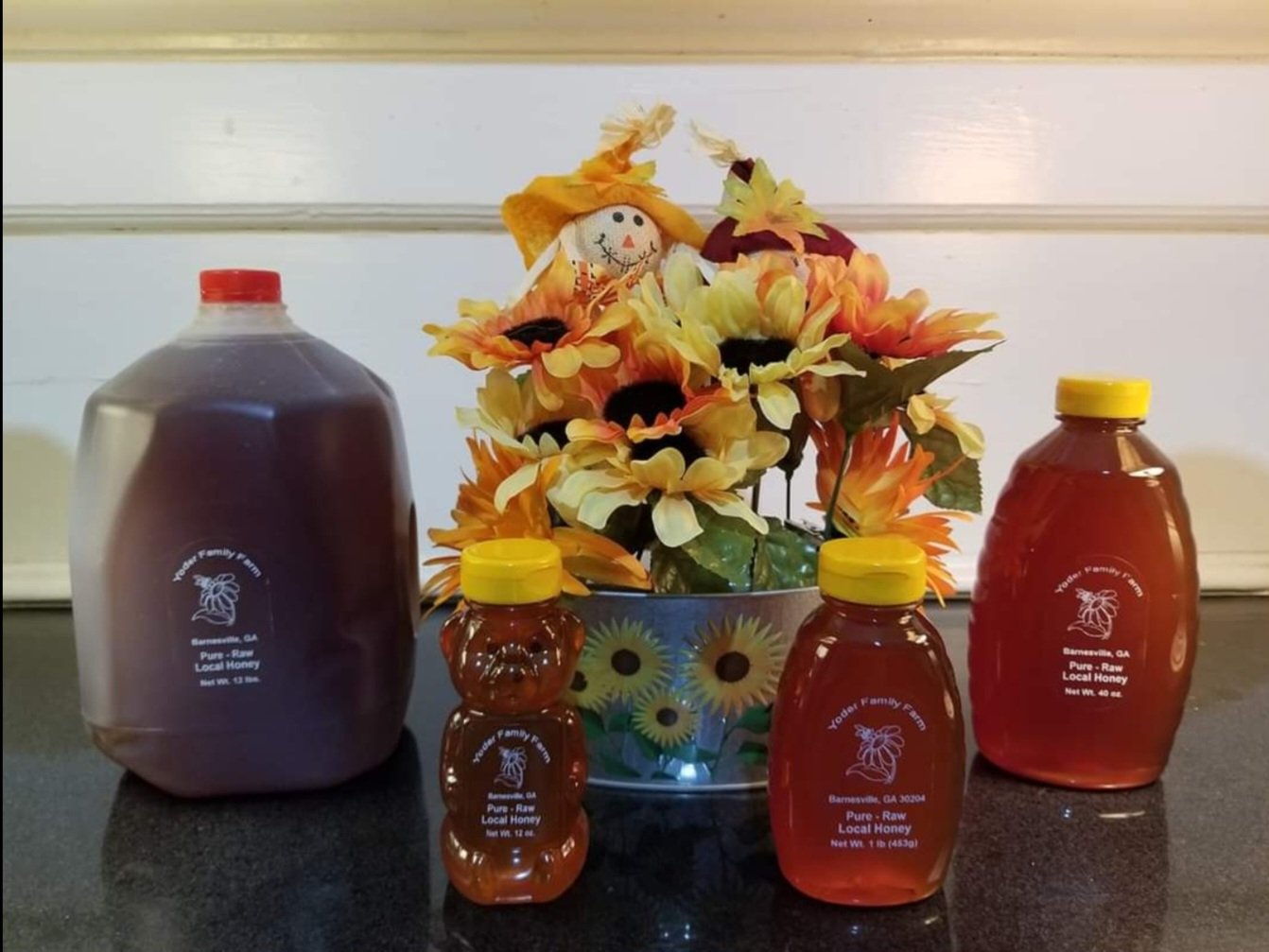 Bottles of pure raw local honey from Yoder Family Farm displayed alongside a vibrant flower arrangement, including sunflowers and a decorative scarecrow, on a dark countertop.